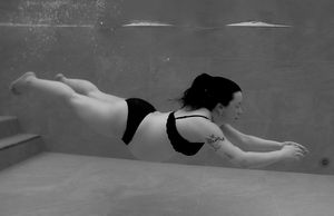 En gravid kvinna i svart bikini svävar under vattnet i en pool. Hon blundar och har sina armar framåt.