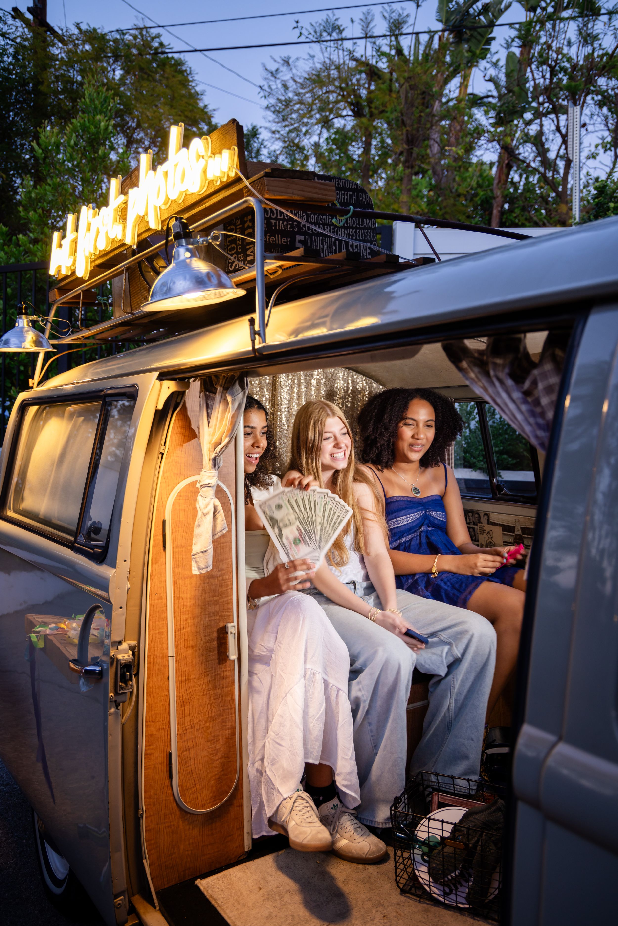 Three young women sitting in a vintage van examining fan of cash at dusk.