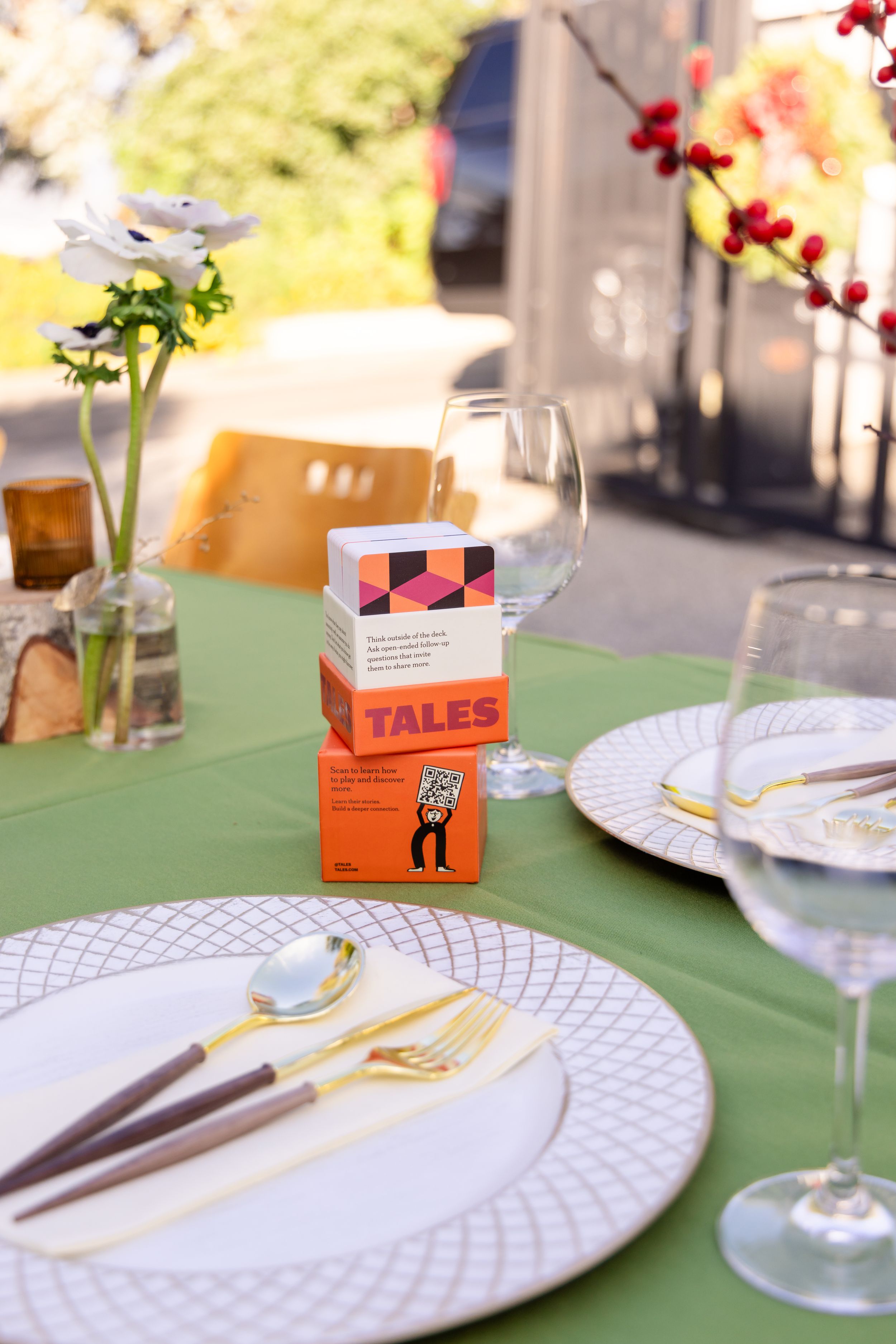 Elegant outdoor table setting with green tablecloth, gold cutlery, and Tales card game centerpiece.