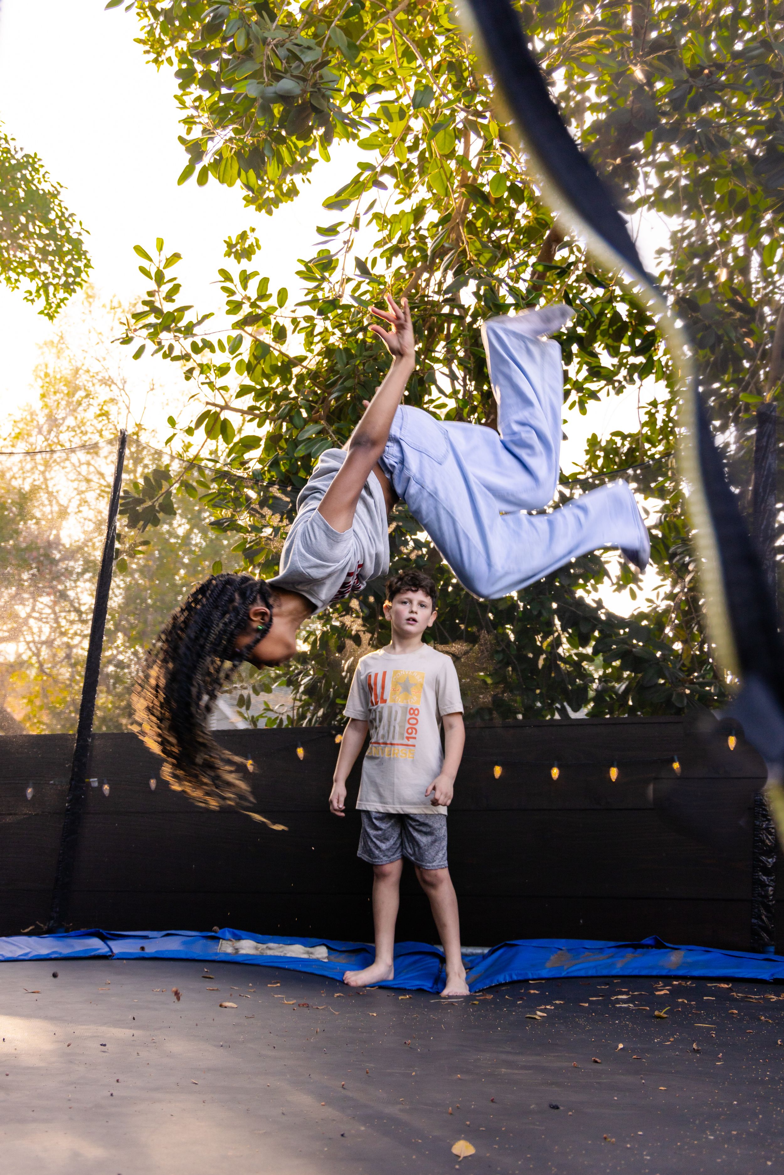 Child doing backflip on trampoline while another child watches in sunlit backyard.