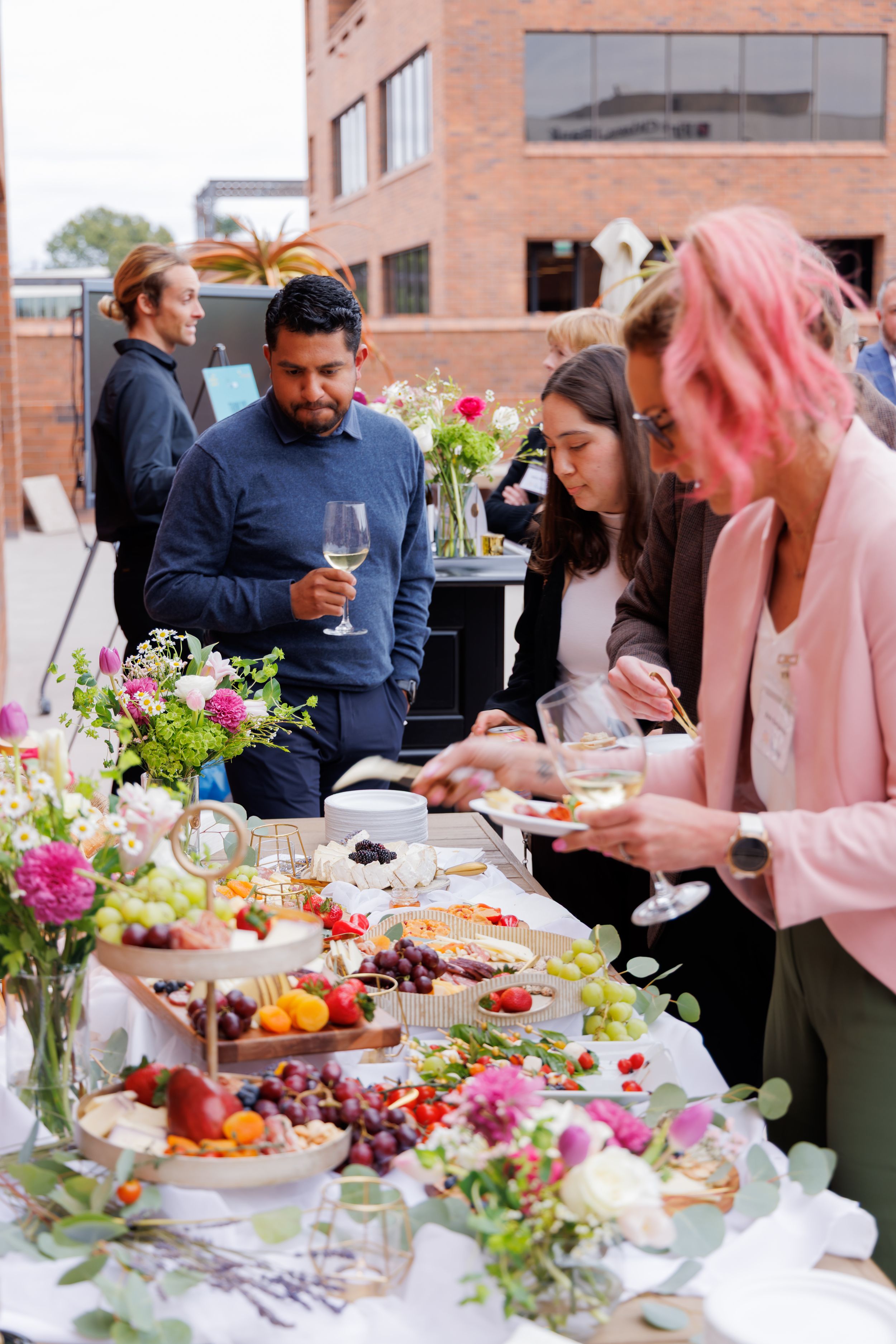 Guests enjoying a colorful outdoor grazing table with fruits, cheeses, and wine at an event.