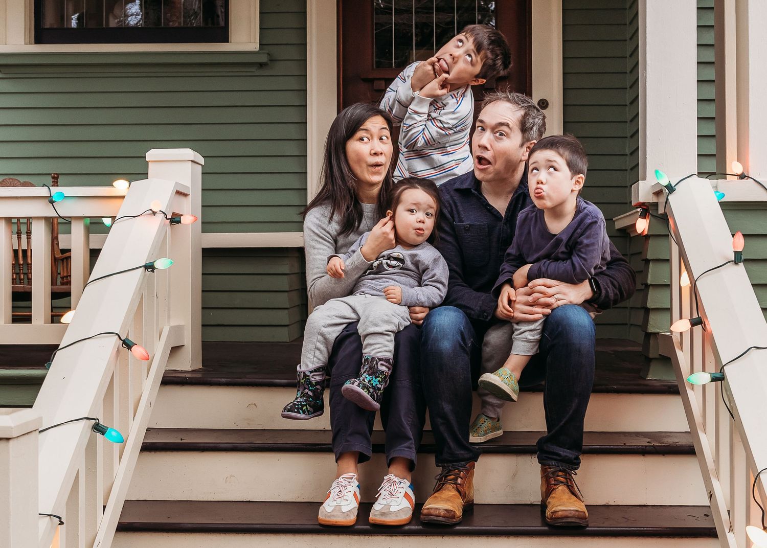 Family gathered on front porch steps of green house decorated with string lights.