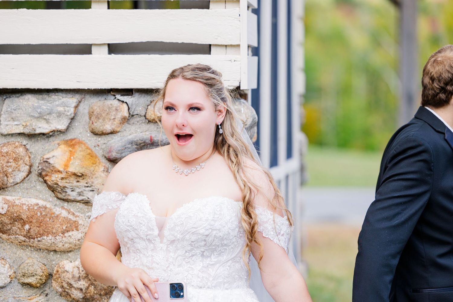 A bride shows excitement while standing near a white slatted wall outdoors.