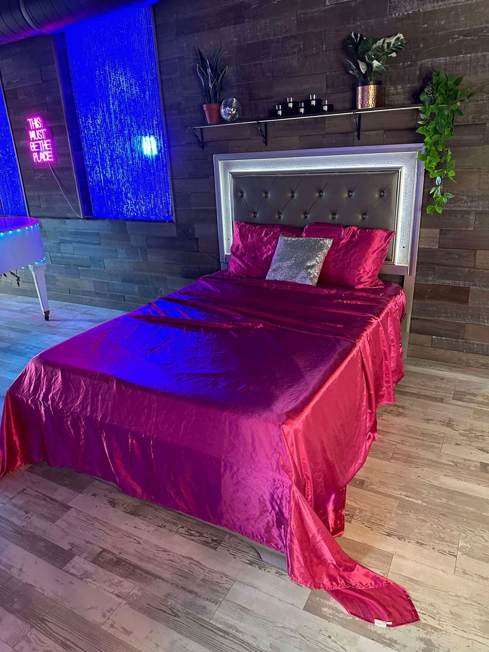 Bedroom setup with red satin bedding and blue neon accent lighting against wooden wall.