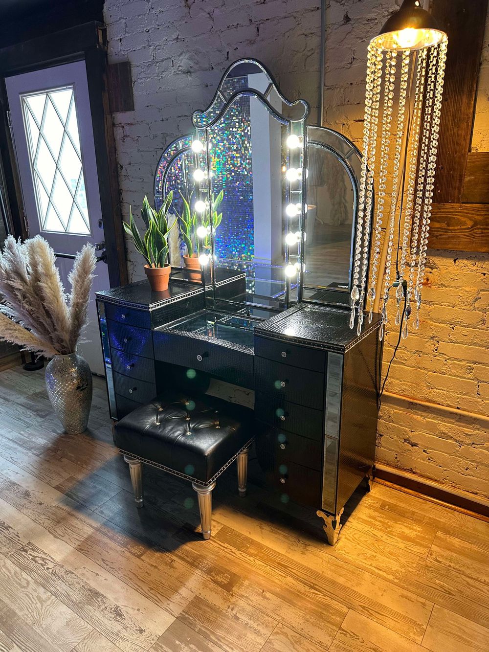 Black makeup vanity with illuminated mirror and crystal chandelier against brick wall.