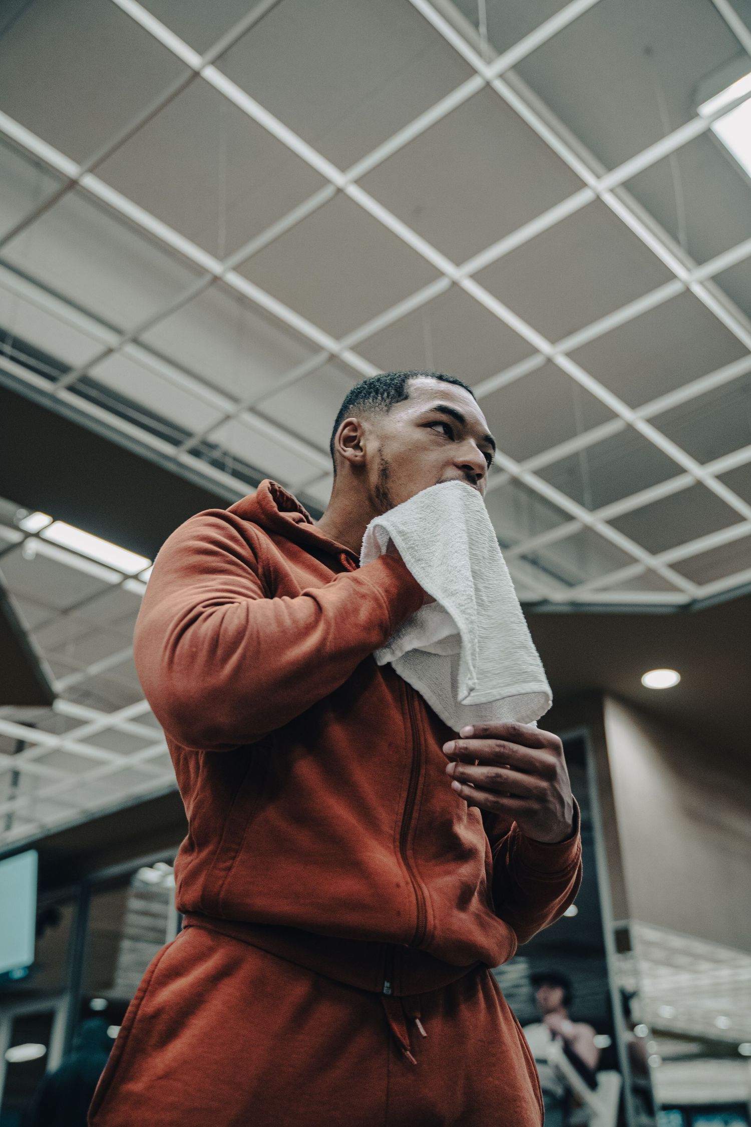 Athletic person in orange hoodie wiping face with towel in gym setting.