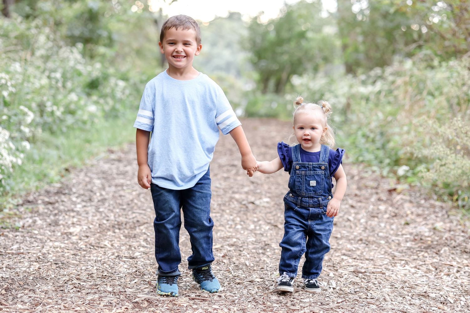 Two siblings hold hands and smile together on a trail surrounded by greenery