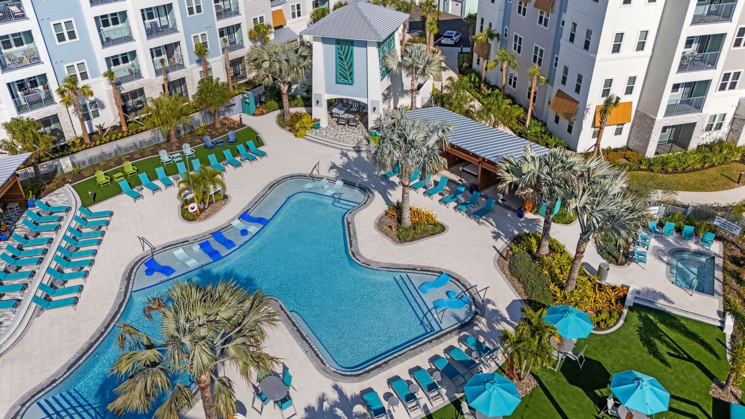 Aerial view of a resort swimming pool with loungers, gazebos and tropical landscaping surrounded by modern buildings.