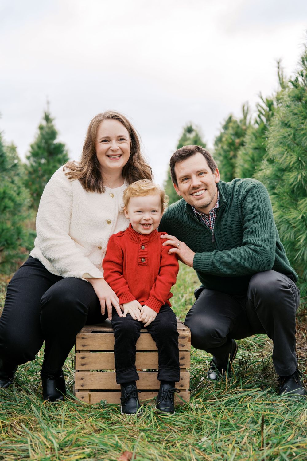 Family portrait on wooden crate surrounded by evergreen trees at Christmas tree farm.