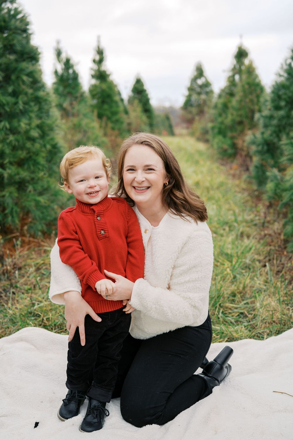 A parent and child share a joyful moment while sitting on a blanket among evergreen trees.