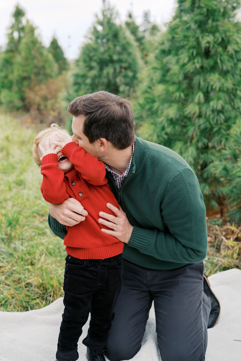 A tender moment between parent and child wearing red and green sweaters among evergreen trees.