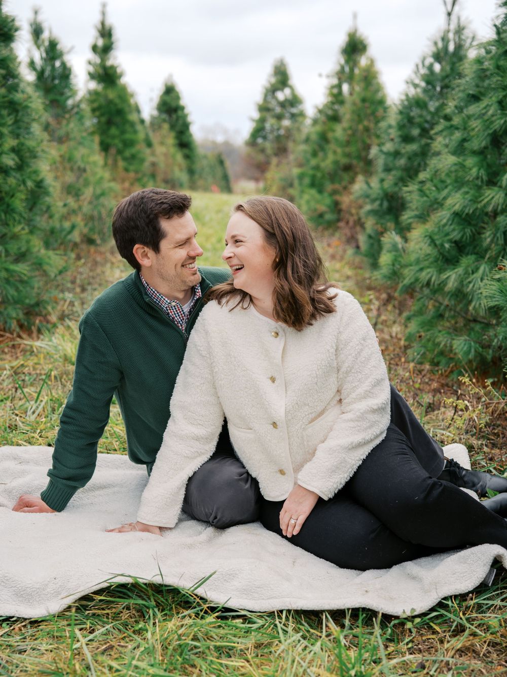 A couple shares a loving laugh while sitting on a blanket surrounded by pine trees.