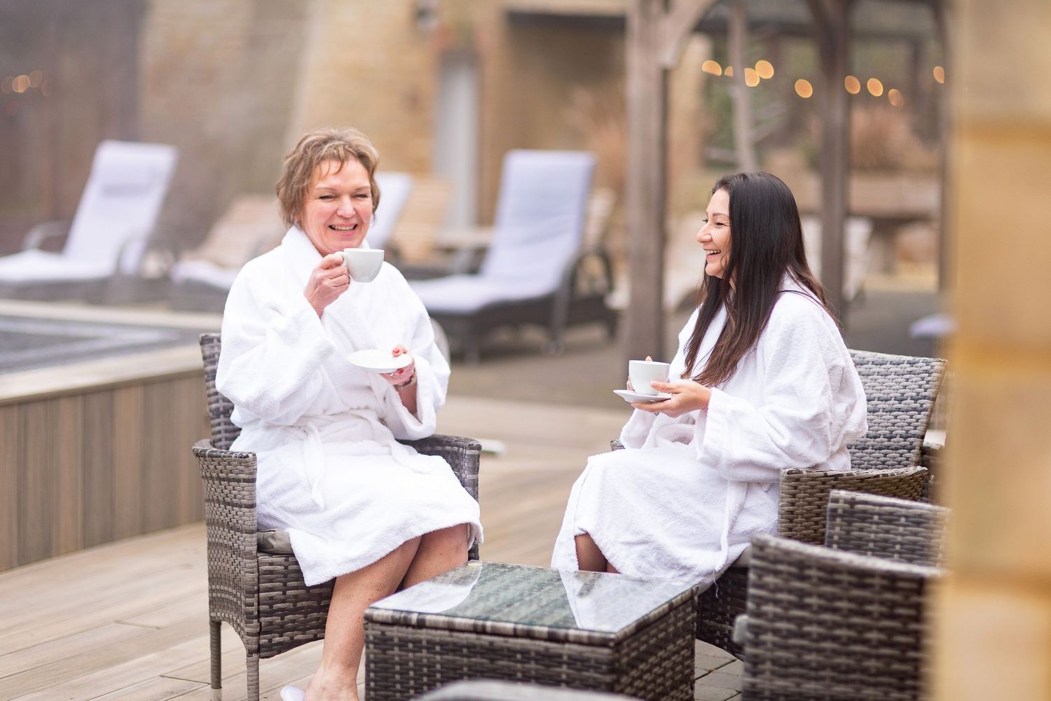 Two people in white robes relaxing in spa chairs on an outdoor deck while having drinks.
