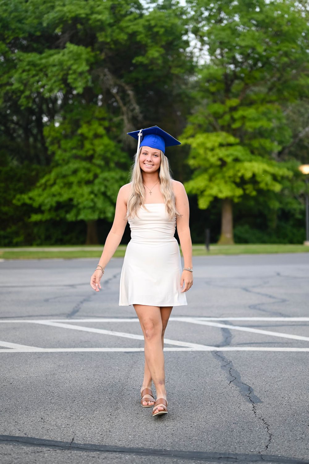 Graduate in white dress and blue mortarboard poses playfully in empty parking lot.