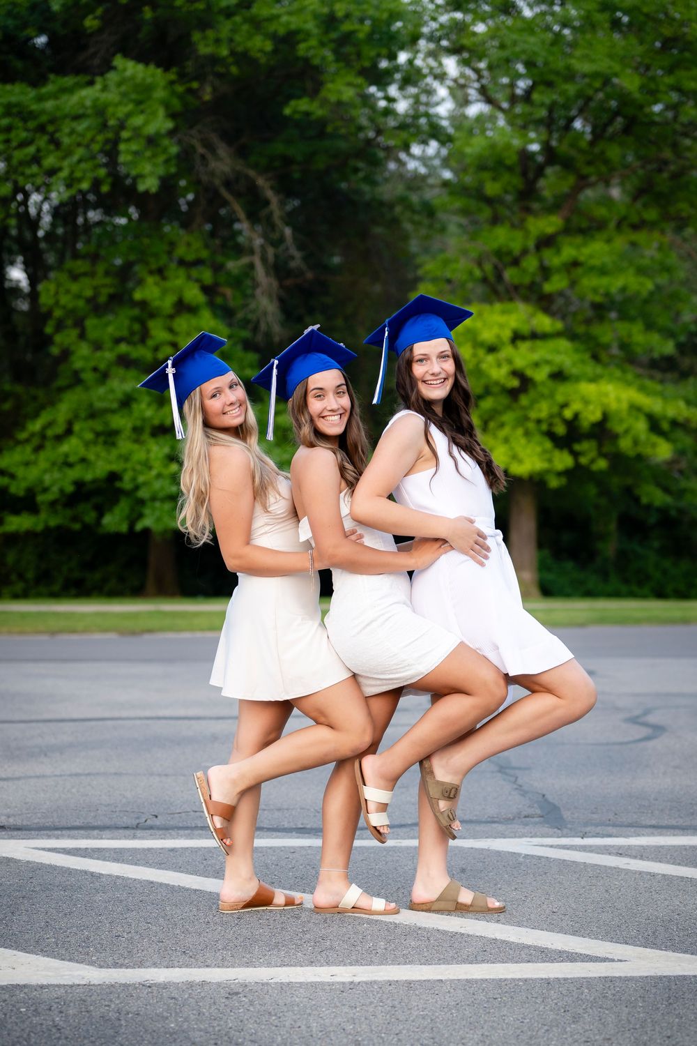 Graduates in white dresses and blue caps pose playfully in a parking lot.