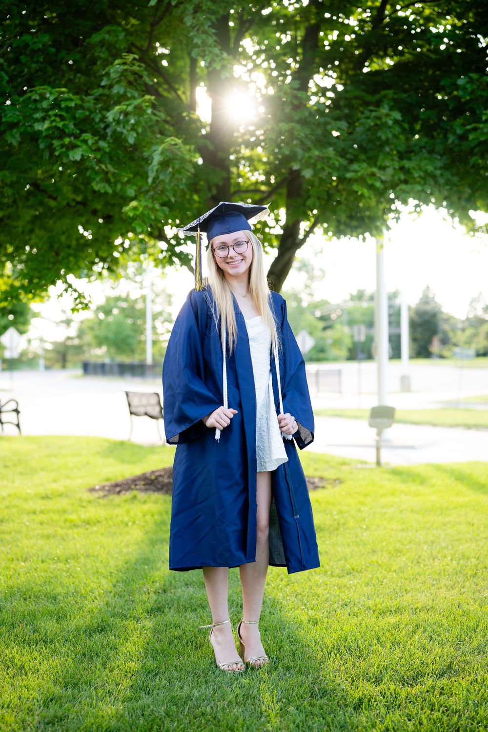 Smiling female graduate in blue cap and gown standing on green grass outdoors.