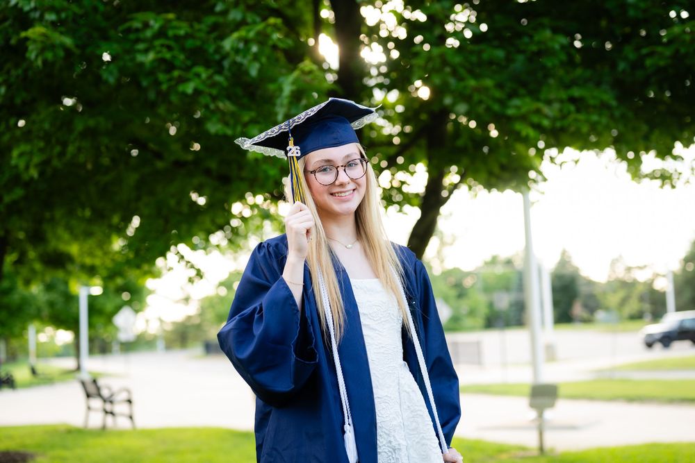 Young woman in blue graduation cap and gown smiling outdoors with green trees in background.