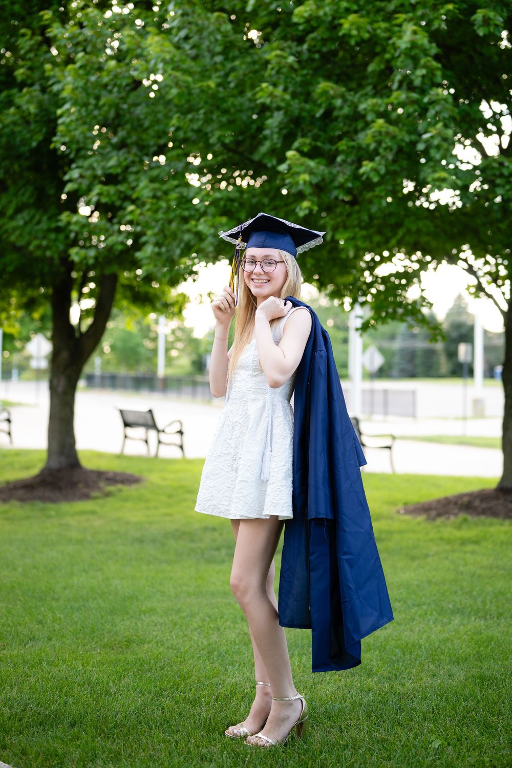 Young woman in white dress and navy graduation cap and gown smiling outdoors on green lawn with trees.