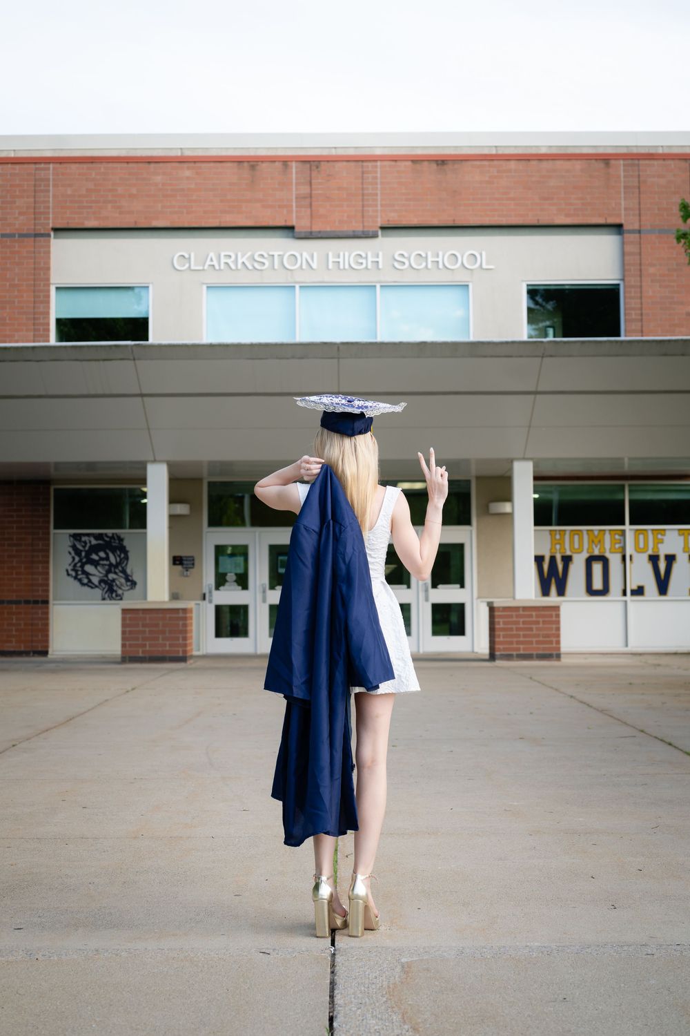A blonde graduate in a white dress holds her navy cap and gown outside Clarkston High School, flashing a peace sign.