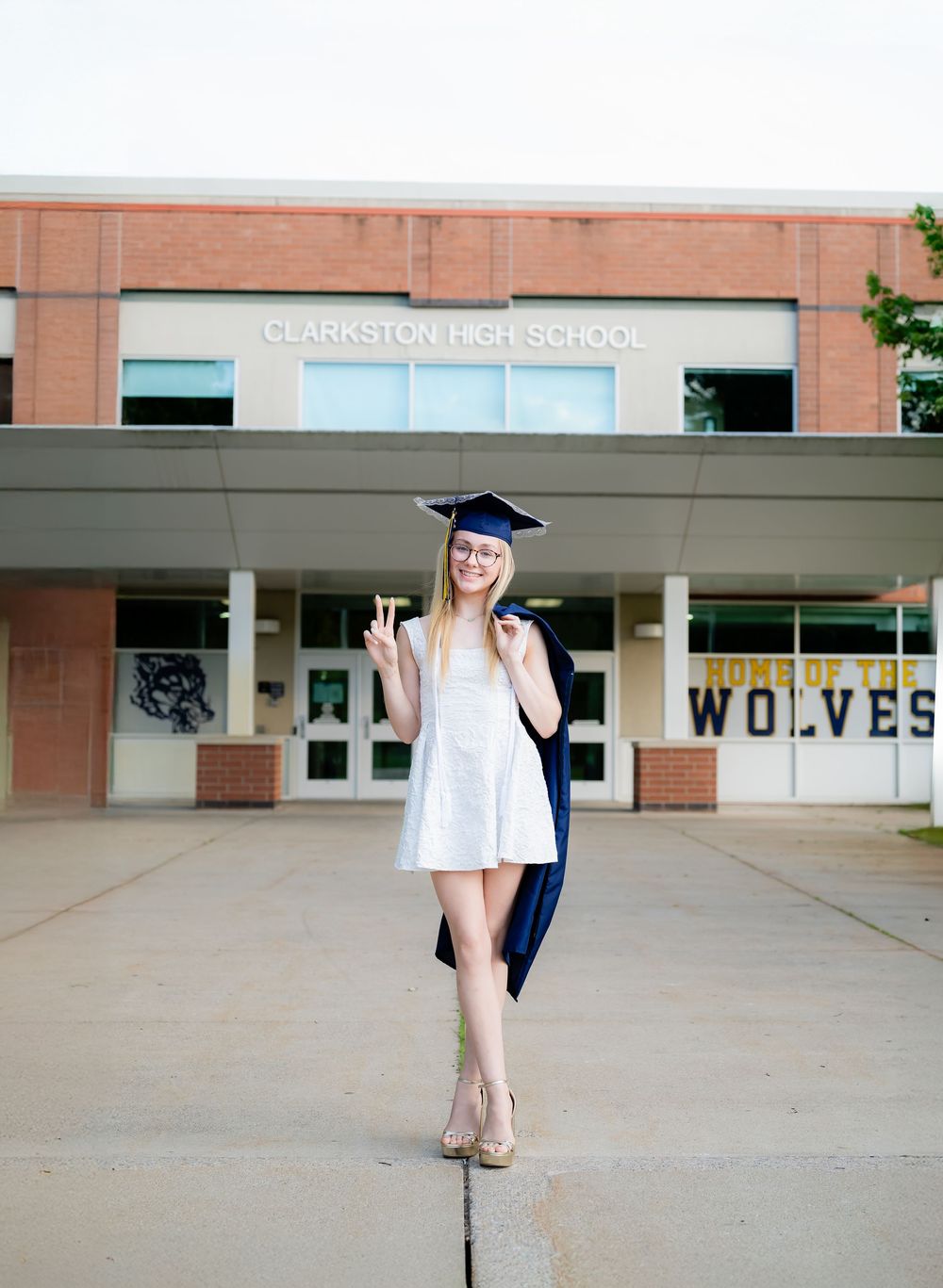 A smiling graduate poses in cap and gown outside Clarkston High School, home of the Wolves.