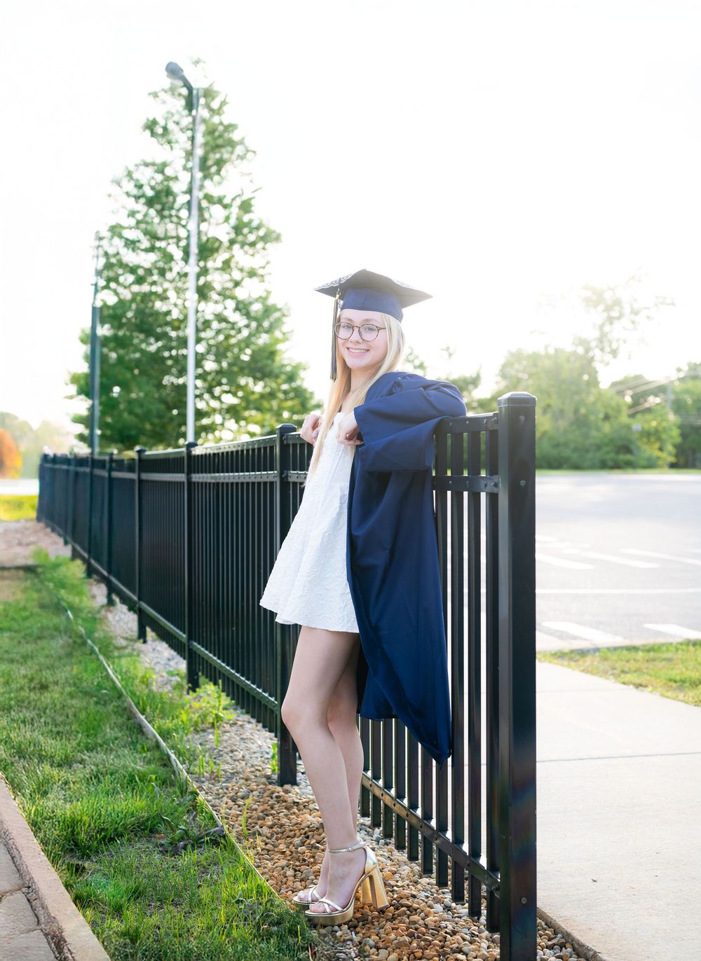 Young woman in navy graduation cap and gown poses by black iron fence outdoors on a sunny day.