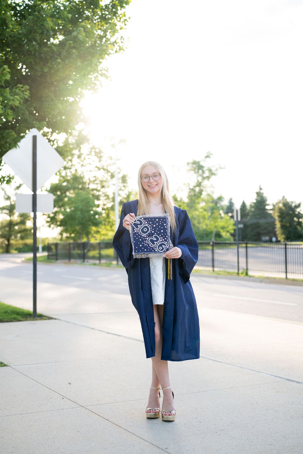 Smiling blonde graduate in navy cap and gown holding diploma on sunny sidewalk outdoors.