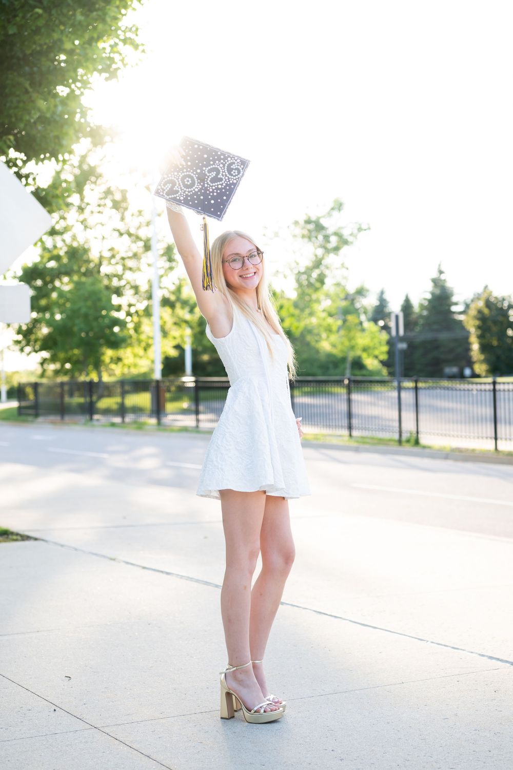 Excited female graduate in white dress raising cap outdoors on sunny day.