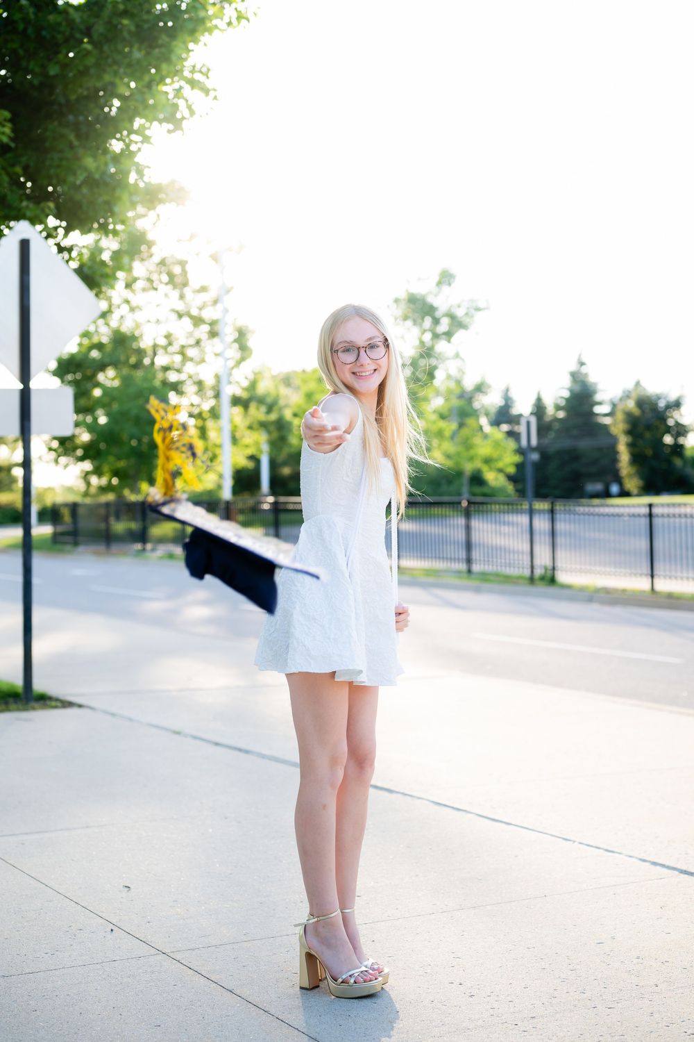 Smiling blonde teen girl in white dress and heels holding graduation cap outdoors on sunny day.