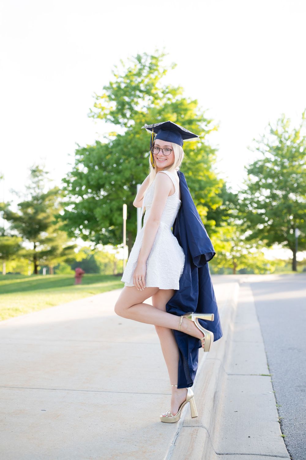 Young woman in white dress, graduation cap and gown, posing outdoors on a sunny day.