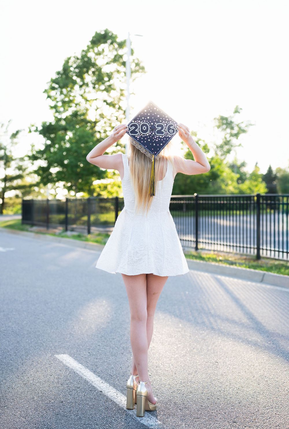 Young woman in white dress holding graduation cap with 2026, standing on outdoor path with trees in background.