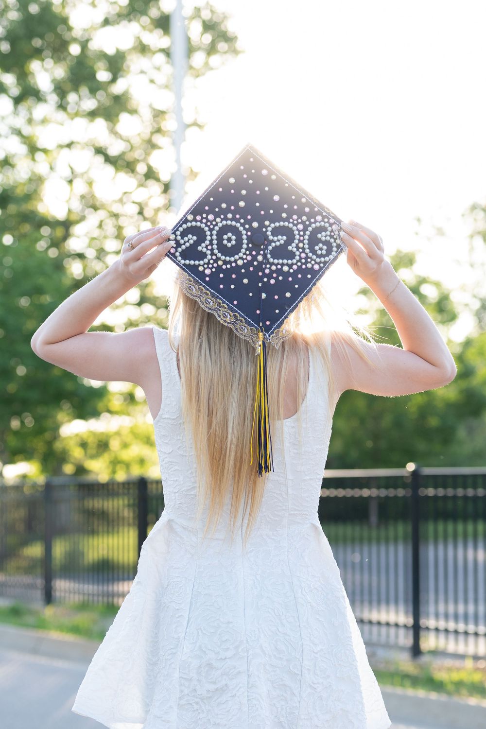 Blonde woman in white dress holding decorated navy blue graduation cap with 2026, viewed from behind outdoors.