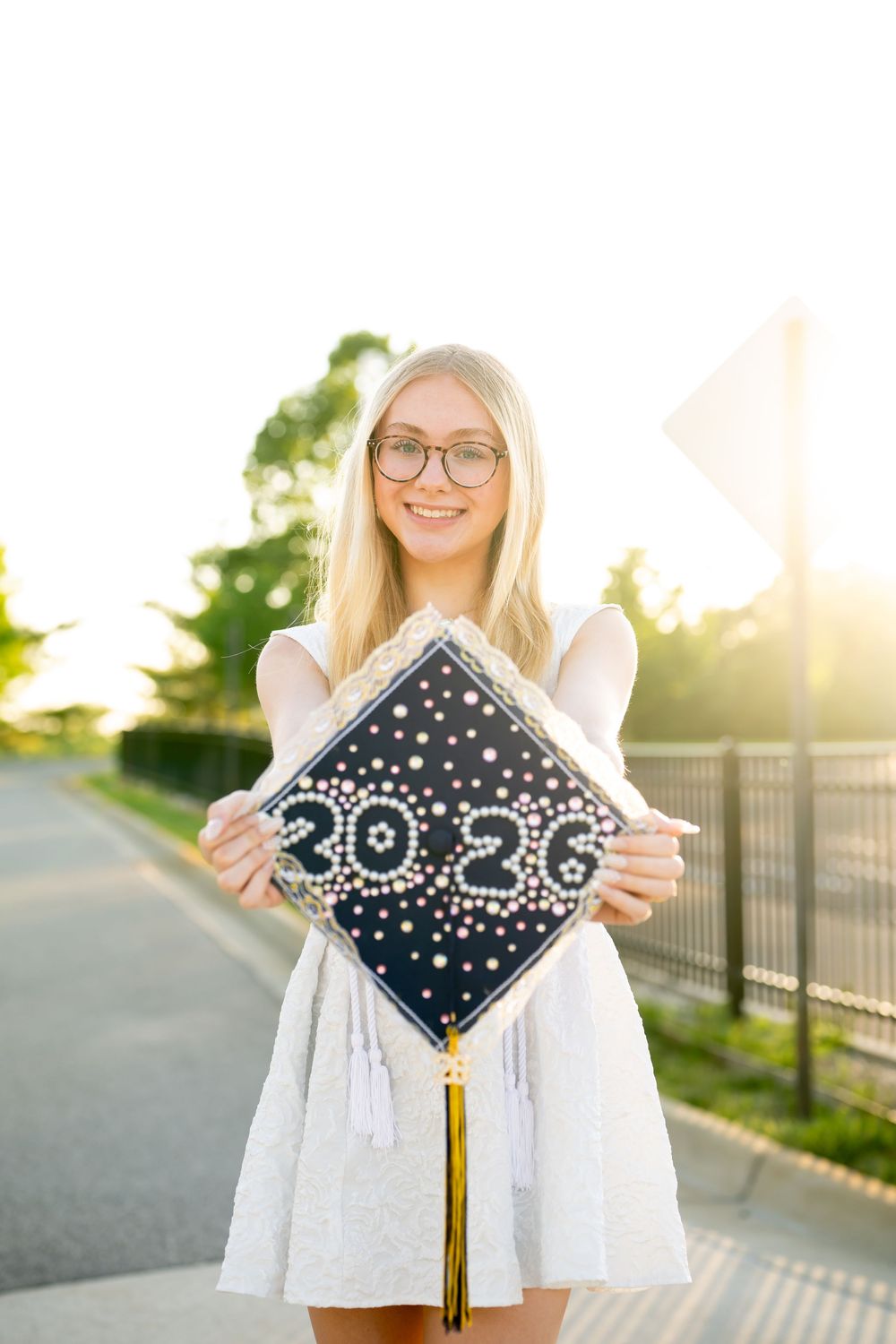 Smiling blonde girl in white dress holding decorated 2026 graduation cap outdoors in golden sunlight.