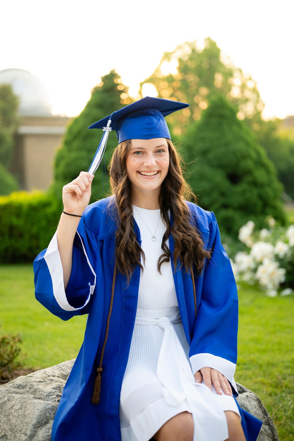 Graduate in blue cap and gown poses on rock during sunset photo session.