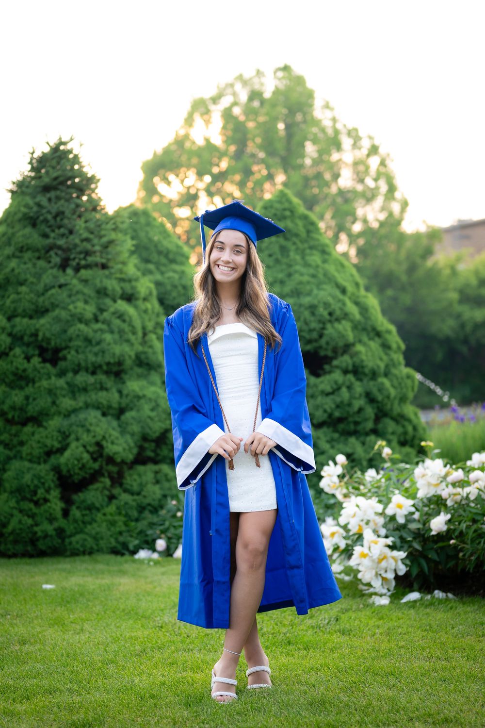 Graduate in blue academic regalia poses playfully in blooming garden setting.