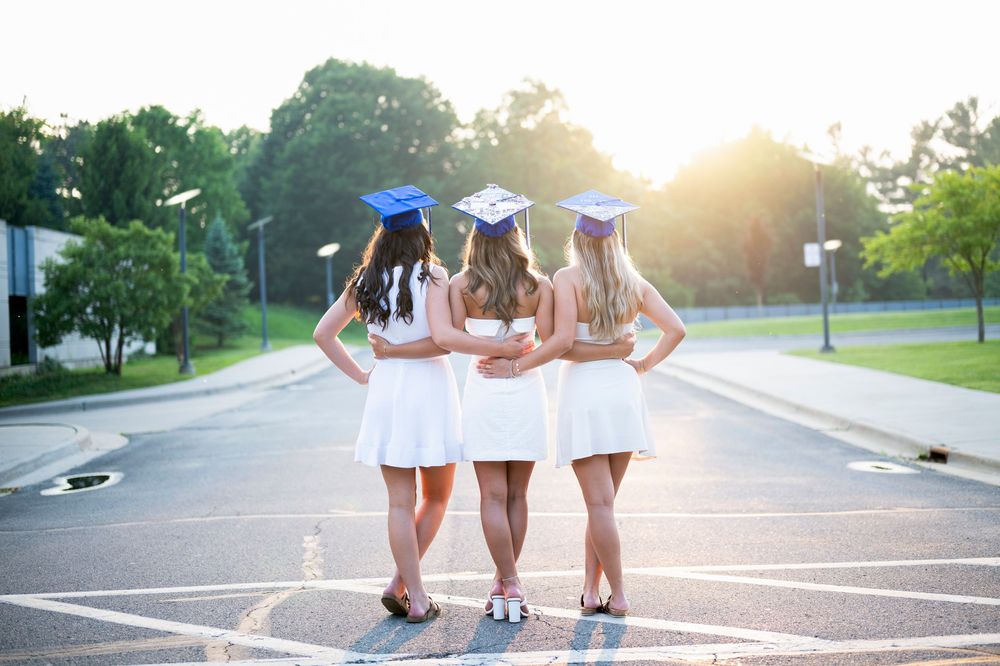 Three graduates in white dresses and blue caps walking together at sunset.