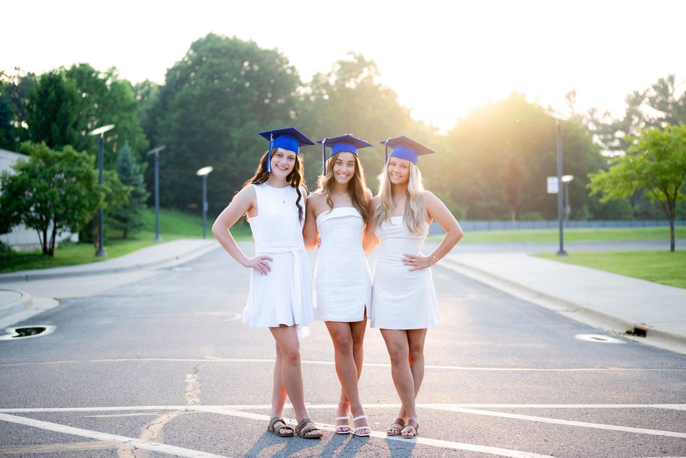 Three graduates in white dresses and blue caps posing together on street.