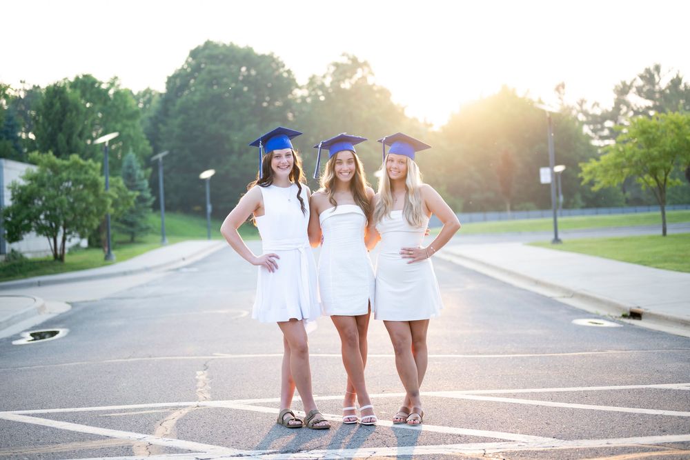 Three graduates in white dresses pose together in a parking lot at sunset.