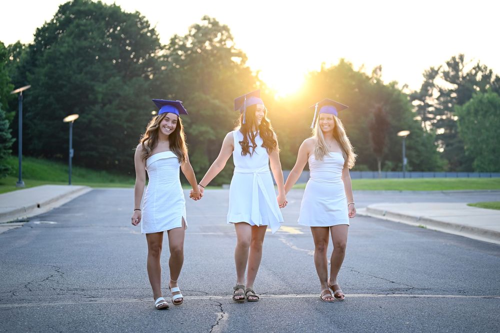 Three graduates in white dresses holding hands while walking at sunset.