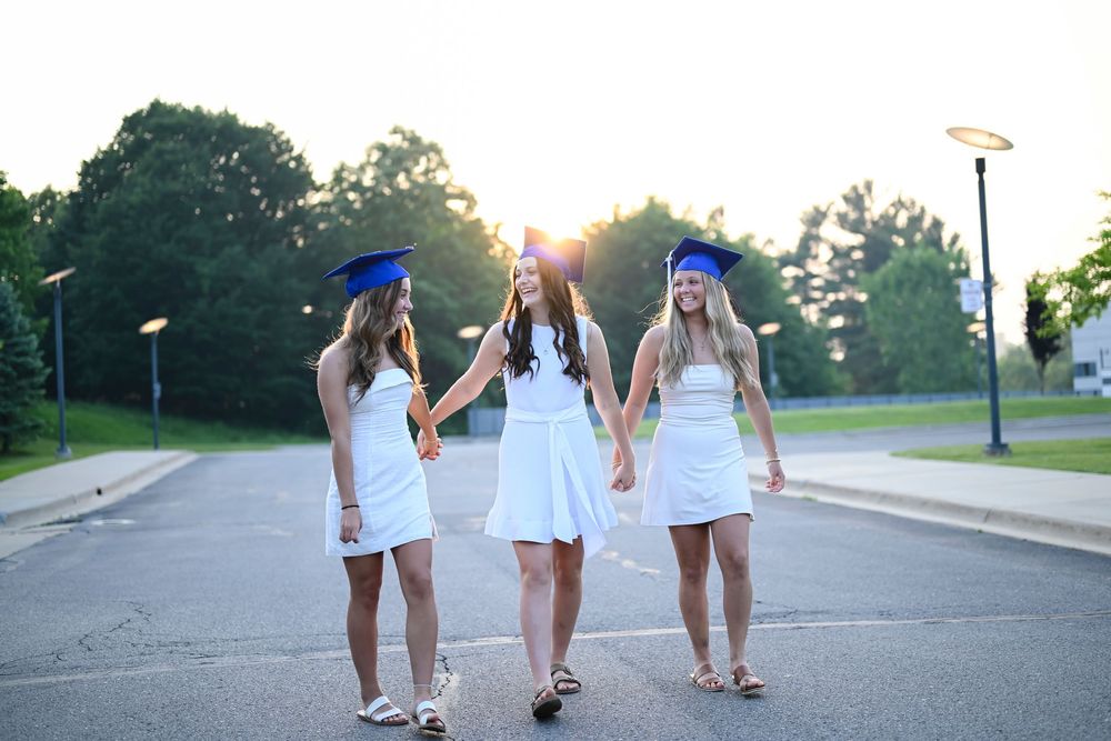 Three graduates in matching white dresses and blue caps walking together outdoors.