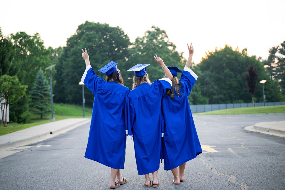 Three graduates in blue gowns raise their arms in celebration outdoors.