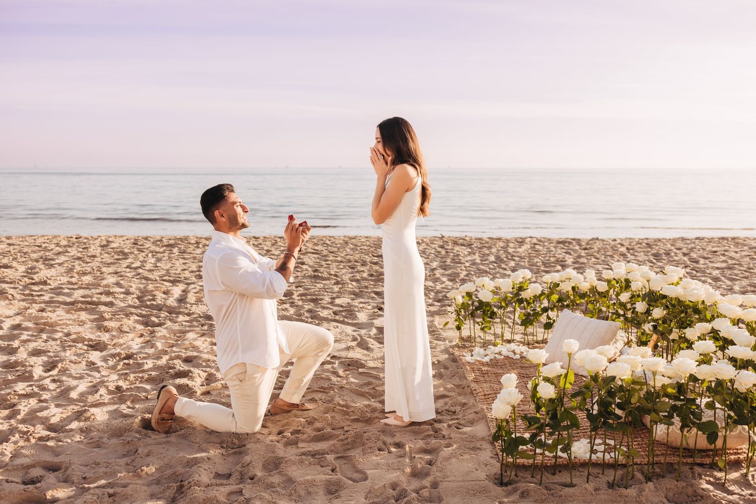 Romantic couple embraces on sandy beach at sunset with beachfront houses in background.