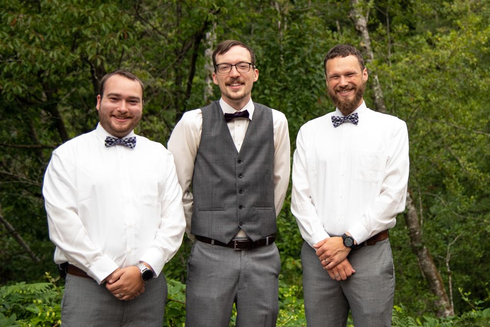 Three men in formal wedding attire pose together against green foliage background.