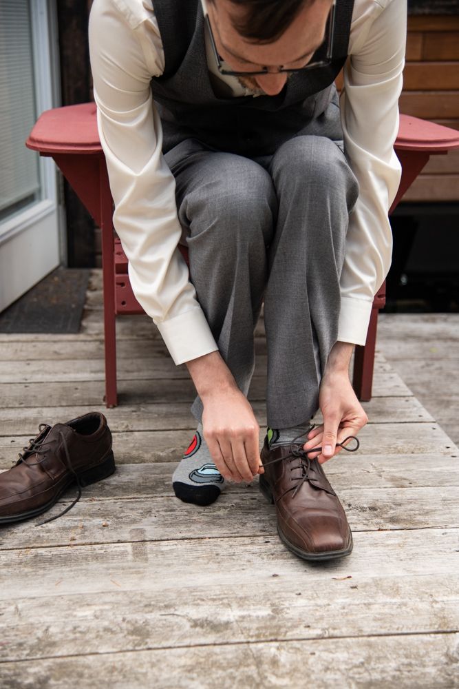 Groom in formal attire putting on brown dress shoes while seated on red chair.