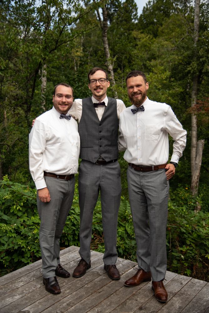 Three groomsmen in formal attire pose together on a wooden deck surrounded by lush green forest.
