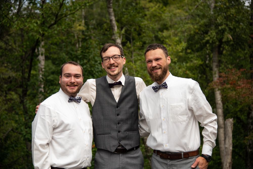 Groomsmen trio in bowties and formal wear pose together in front of trees during wedding photos.