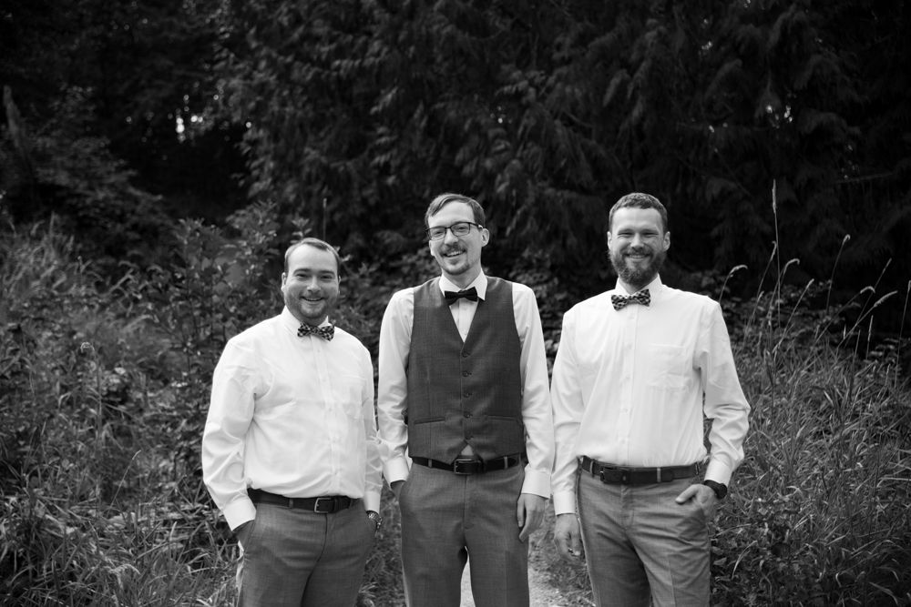 Black and white photo of three groomsmen in formal wear posing against a natural outdoor backdrop.