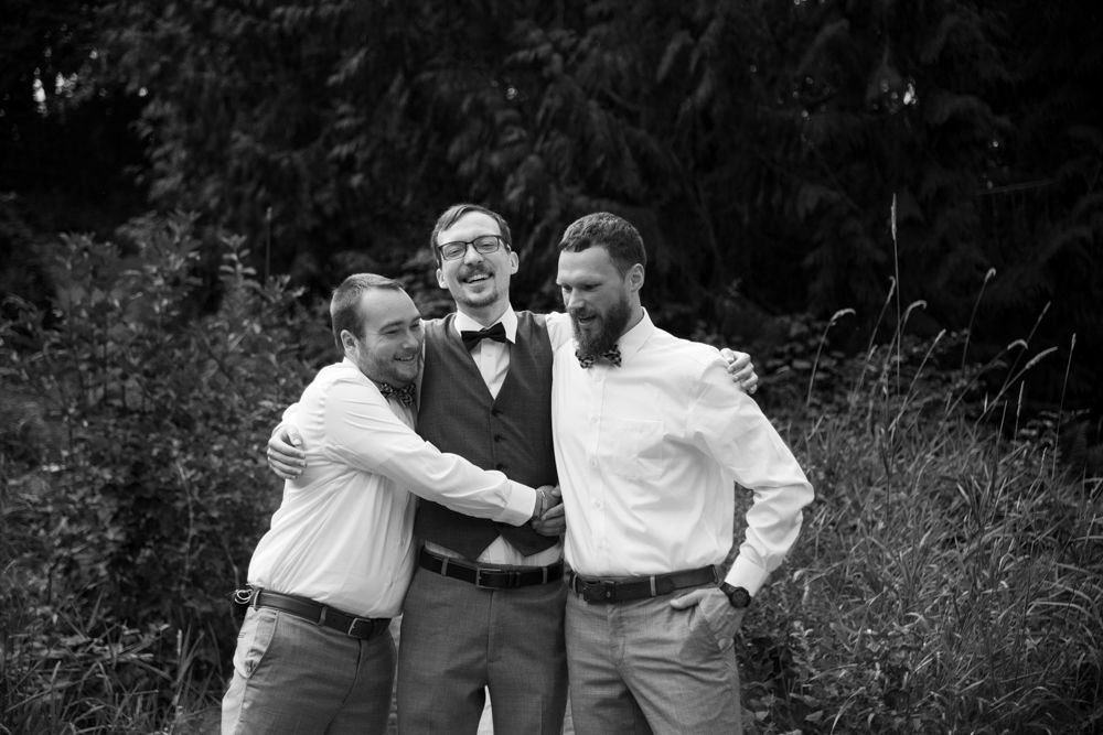 Candid black and white photo of groomsmen sharing a lighthearted moment outdoors.