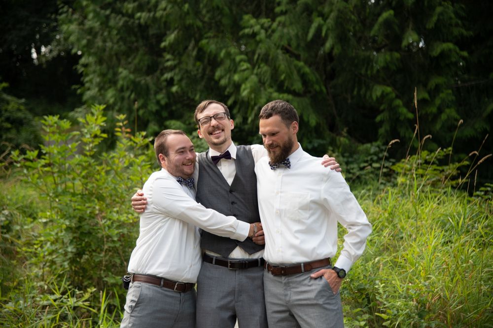 Three groomsmen embrace in a casual group pose surrounded by greenery during wedding photos.