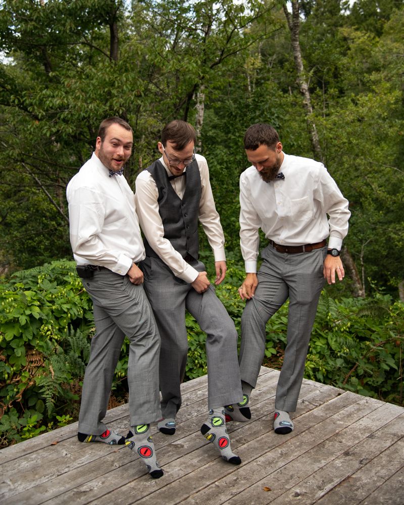 Three people in formal attire showing off their colorful patterned socks while standing on a wooden deck outdoors.