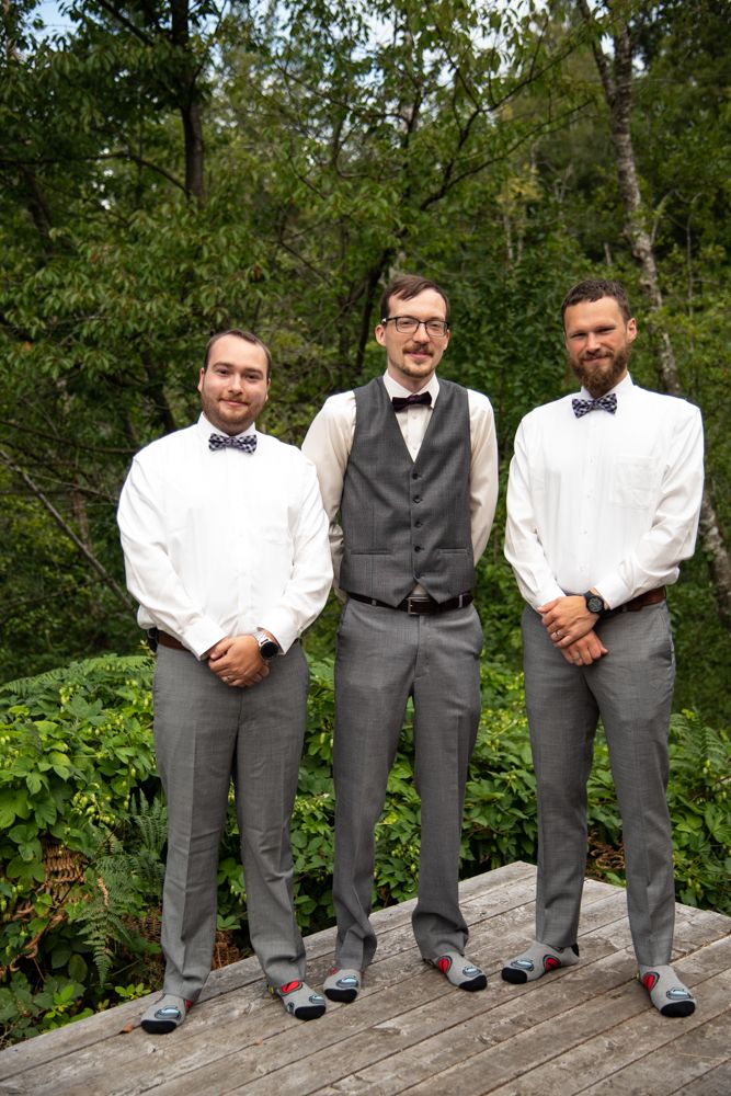 Three groomsmen in formal attire posing together on wooden deck in outdoor setting.